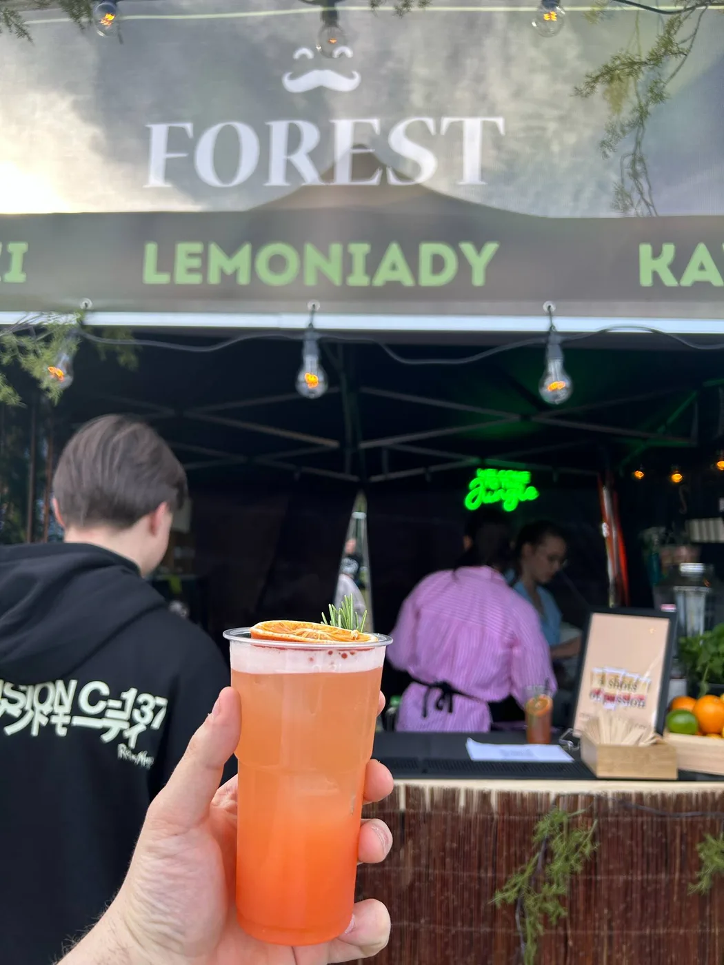 Drink w dłoni na tle stoiska Forest z neonem
